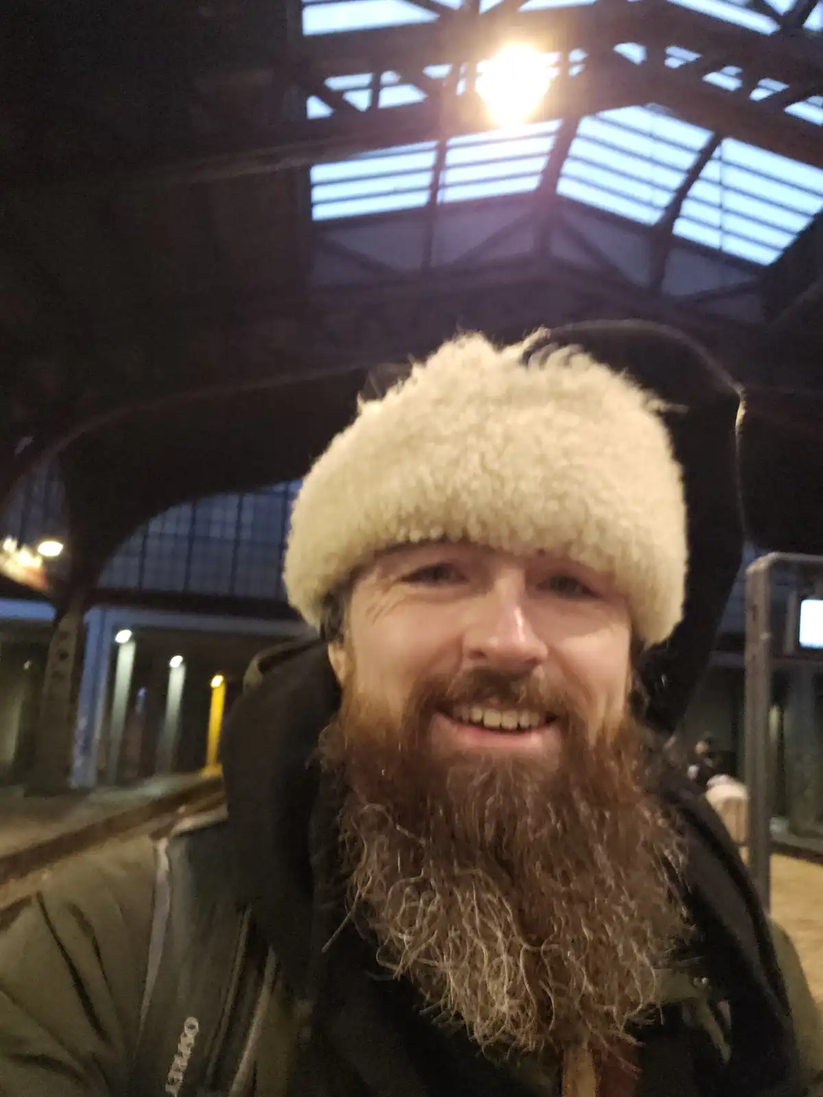 A man with a long, full beard and a fluffy white winter hat smiles at the camera while standing in what appears to be a…
