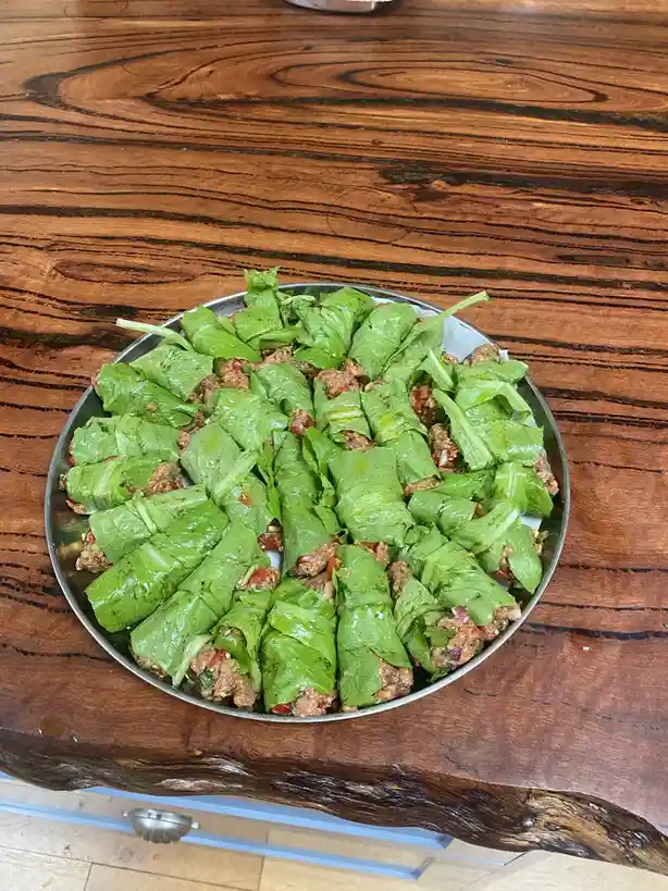 A metal tray holds dozens of lettuce leaves rolled around a meat and vegetable filling, arranged neatly on a rustic wooden…