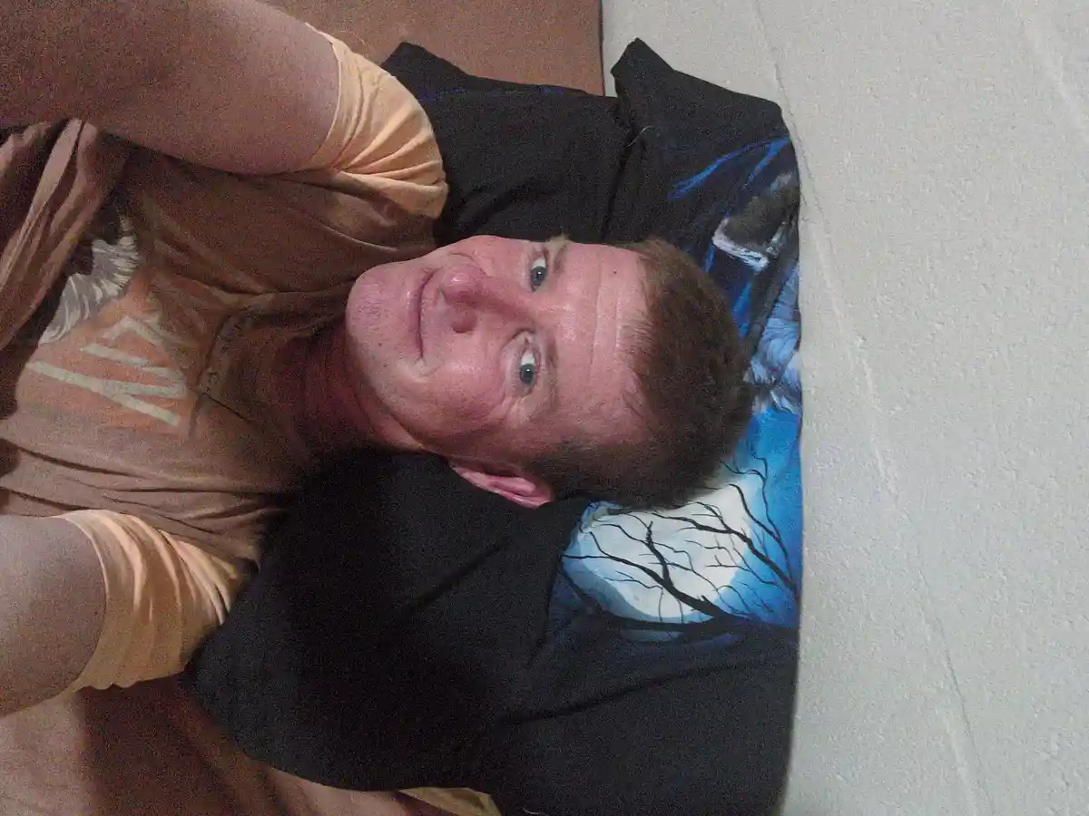 A man with reddish hair and blue eyes lies on his back on a bed with a black and blue pillow featuring a tree design…