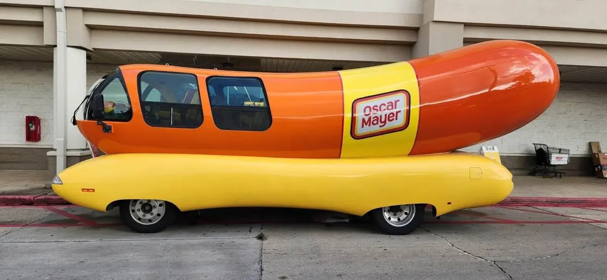 A side view of the iconic Oscar Mayer Wienermobile, a giant hot dog-shaped vehicle with orange and yellow coloring, parked…