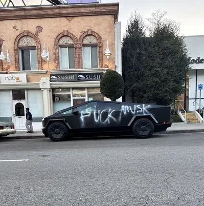 A black Tesla Cybertruck is parked on a city street with the words 'FUCK MUSK' spray-painted in large white letters across…