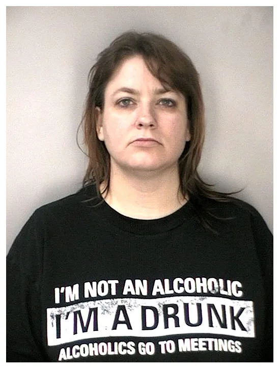 A mugshot-style photo of a woman with brown hair wearing a black t-shirt that reads 'I'M NOT AN ALCOHOLIC I'M A DRUNK…