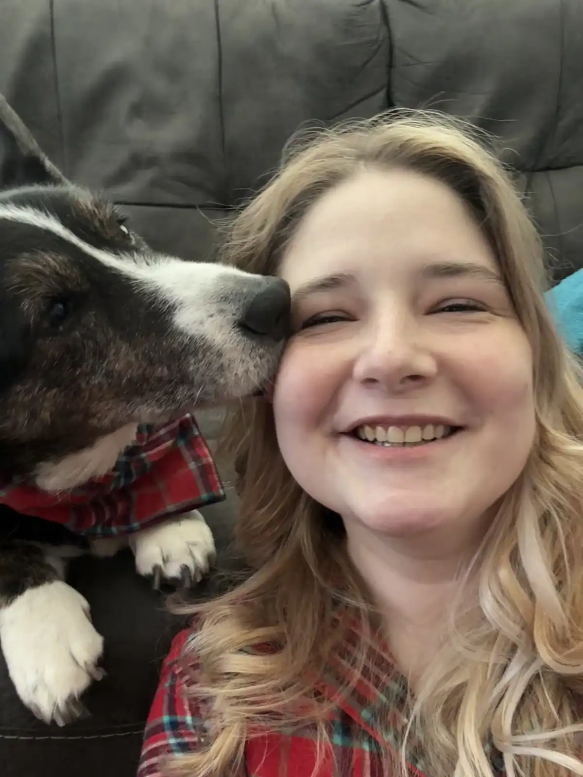 A young woman with blonde hair smiles warmly while a black-and-white dog wearing a red plaid bandana nuzzles her cheek. Both…