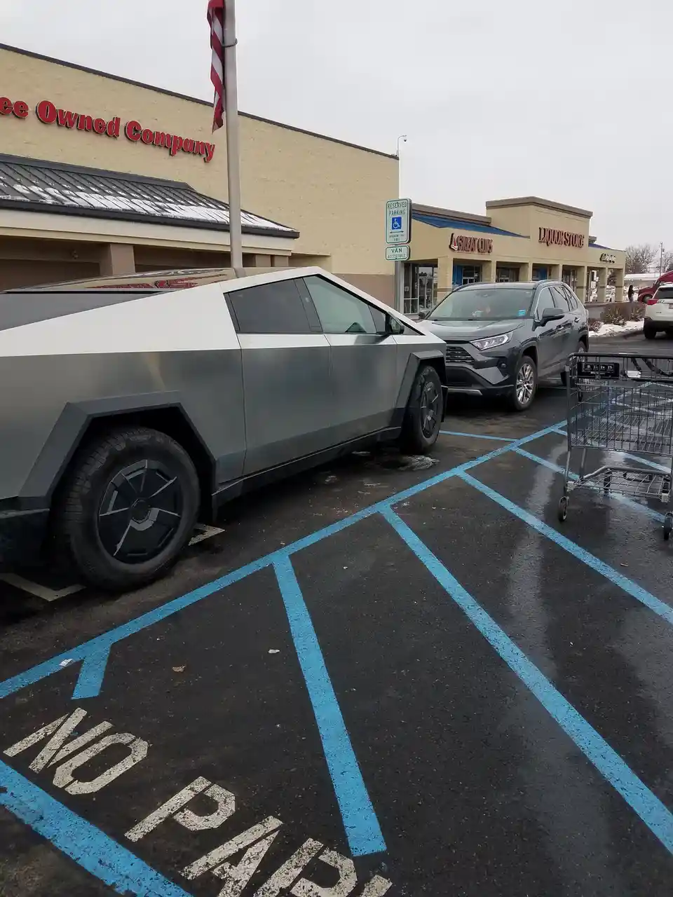 A silver and white Tesla Cybertruck is parked in a blue-striped 'NO PARKING' space in a supermarket parking lot, next to a…