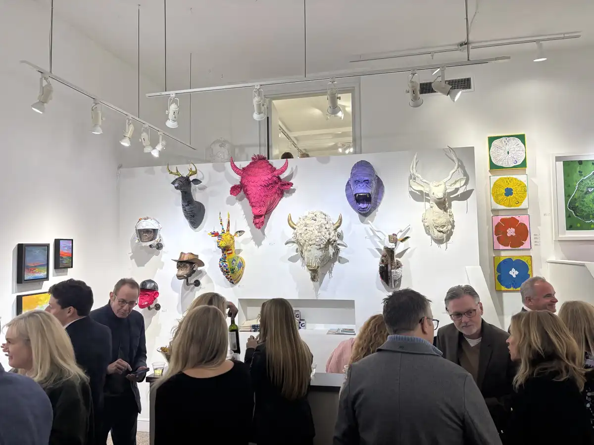 A crowded art gallery features a wall of colorful, stylized animal head sculptures made from unconventional materials like…