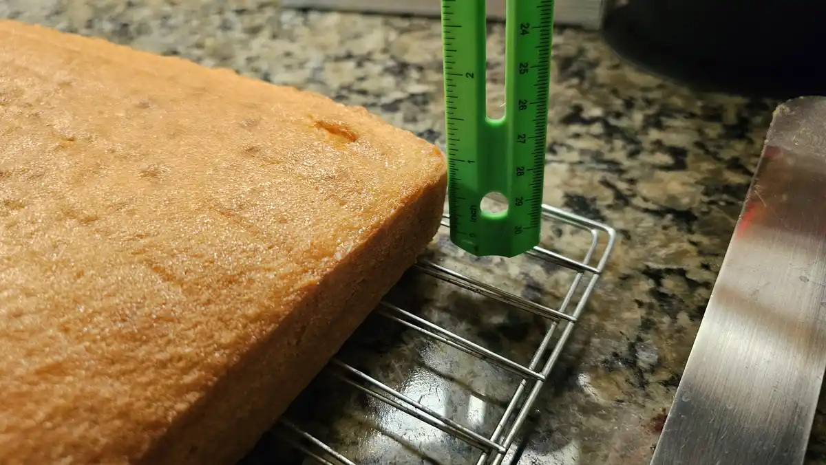A freshly baked golden-brown rectangular cake sits on a metal cooling rack on a granite countertop, with a green plastic…
