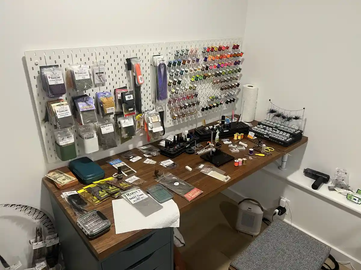 A well-organized fly tying workspace features a wooden desk cluttered with materials like feathers, thread spools, and…