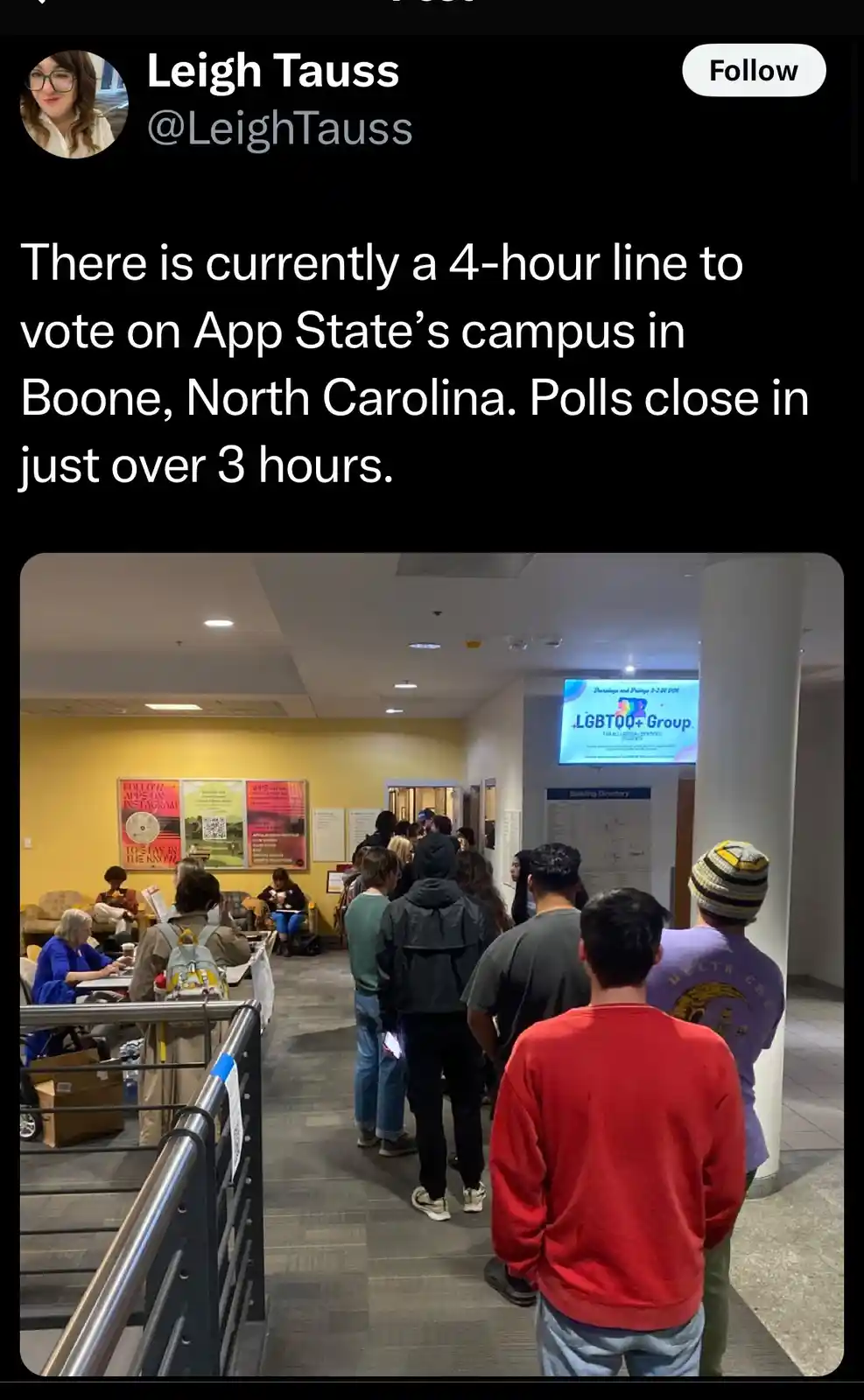 A photo shows a long line of people waiting indoors in a campus hallway, with a digital sign for an LGBTQ+ Group visible in…