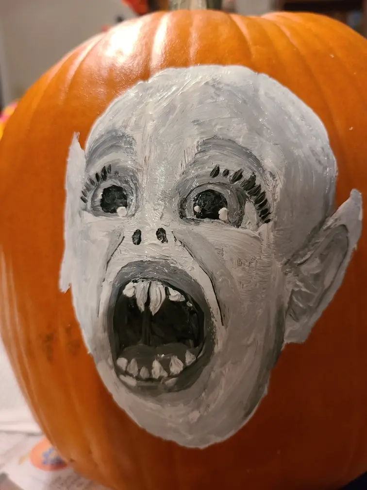 A close-up of an orange pumpkin with a hand-painted face showing wide, black eyes with long lashes, a gaping mouth with…