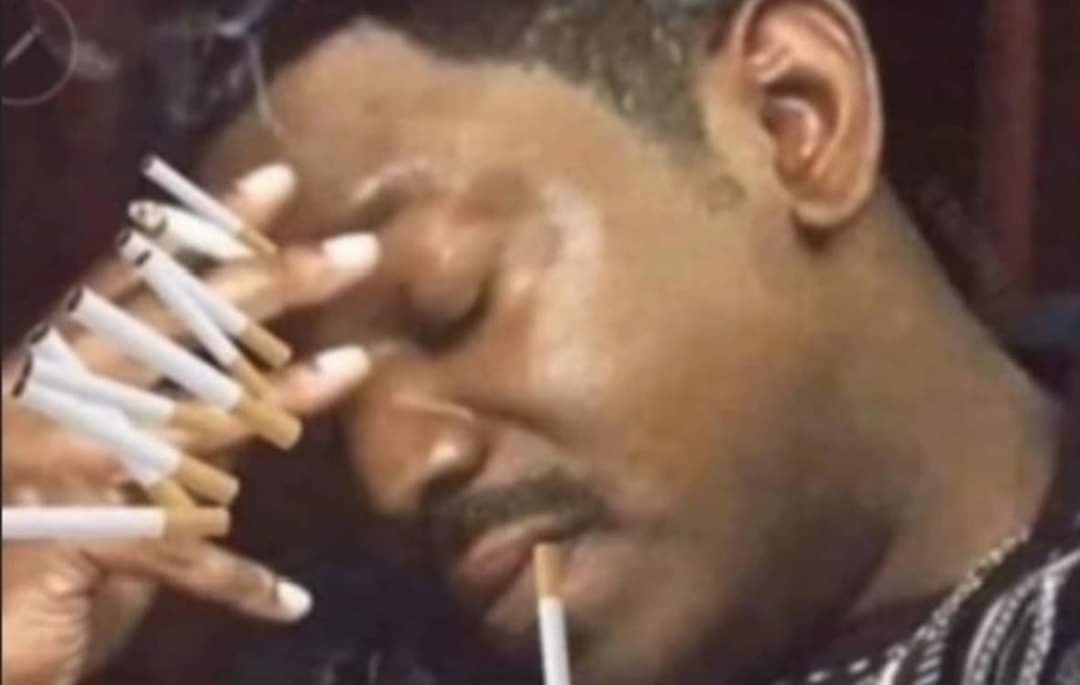 a close-up of a man with his eyes closed, holding multiple cigarettes in his hand near his face, with one cigarette in his…