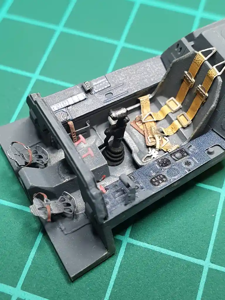 A close-up photo of a detailed, weathered model airplane cockpit with a seat, harness, stick, and pedals, resting on a green…