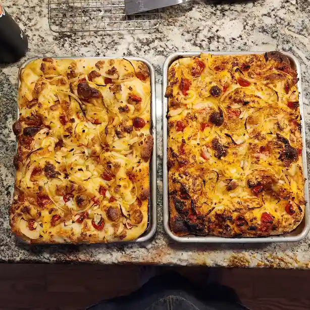 Two rectangular pans of baked pizza sit side-by-side on a granite countertop, both topped with cheese, onions, and cherry…