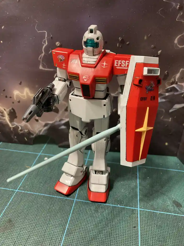 A red and white Gundam model stands on a green grid mat, holding a gun in one hand and a shield in the other, with a…