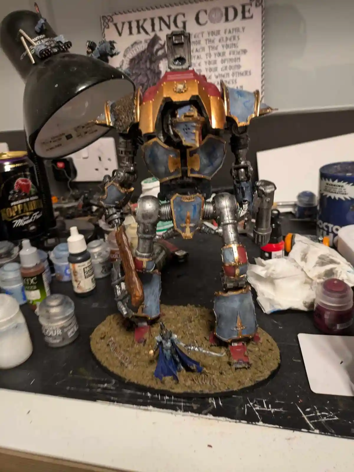 A large, heavily armored, blue-and-gold painted war machine stands on a cluttered hobbyist's desk surrounded by paint…