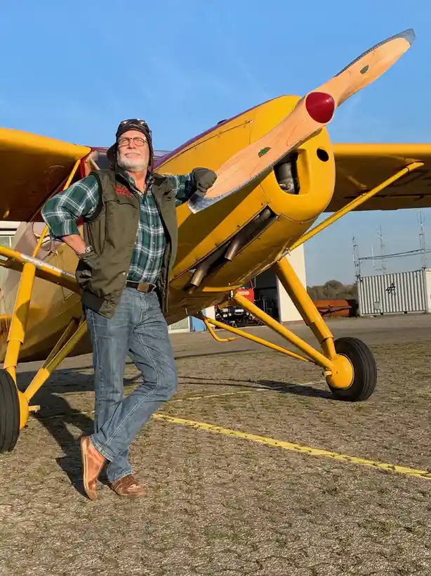 A man with a white beard, glasses, and a pilot's cap stands confidently next to a bright yellow single-engine airplane on a…