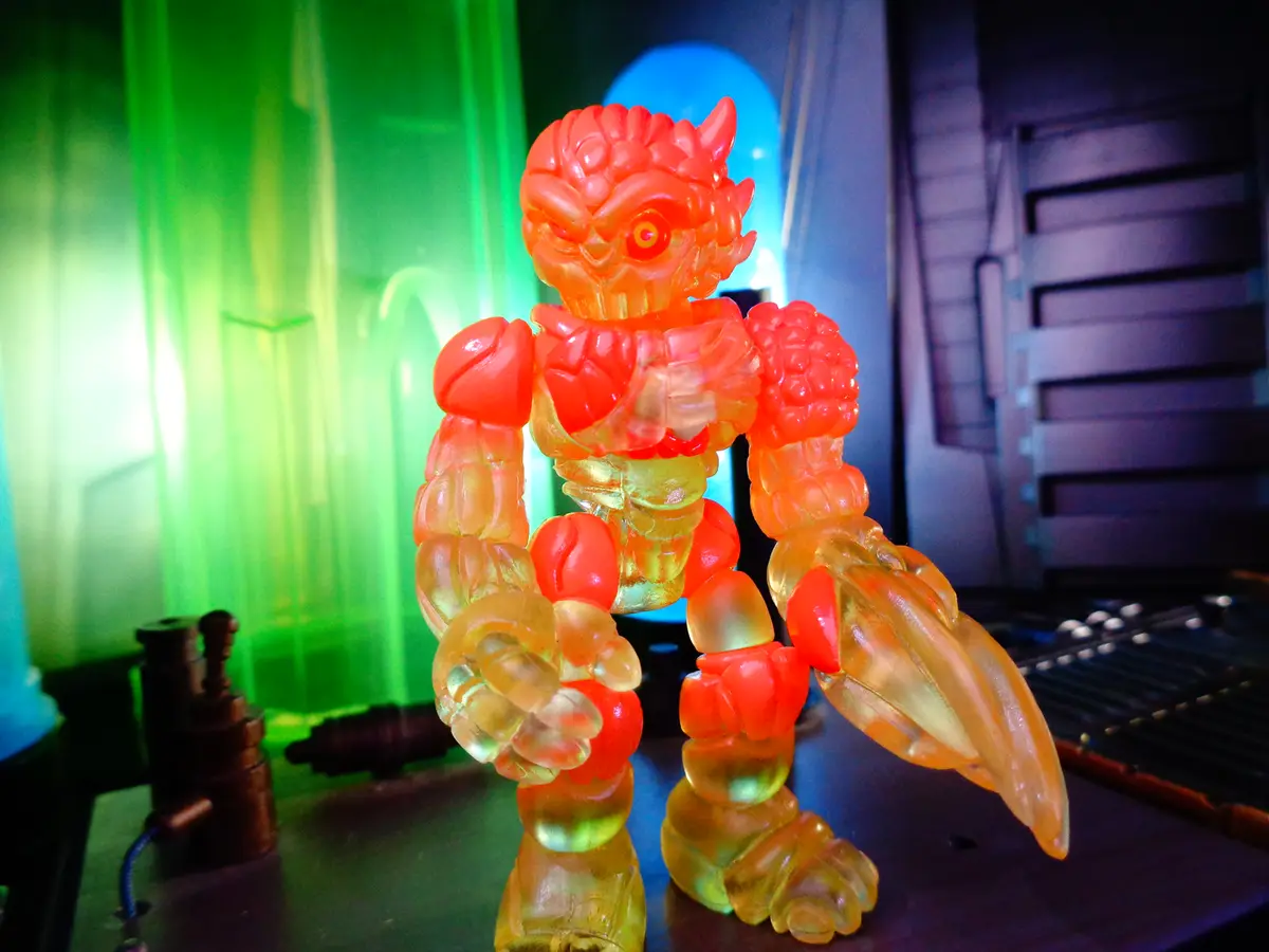 A translucent, orange-red plastic action figure with a demonic face and clawed hands stands in a sci-fi setting with green…
