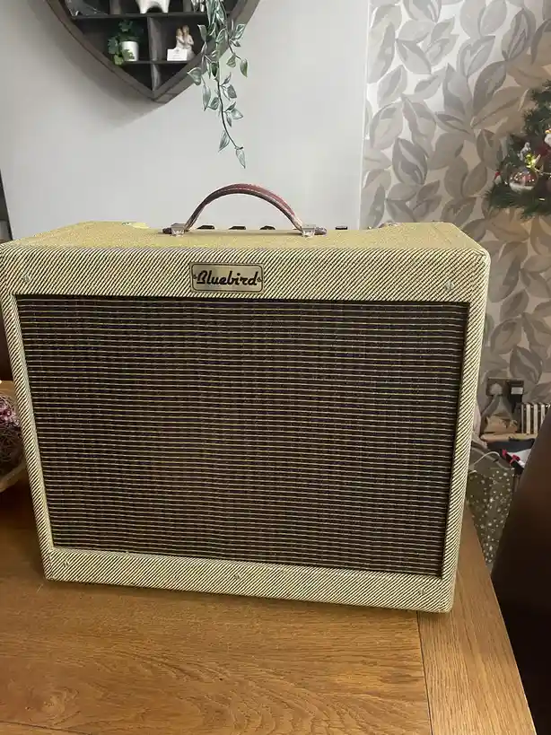 A vintage-style guitar amplifier with a tan tweed exterior and brown grille cloth sits on a wooden table. The brand name…