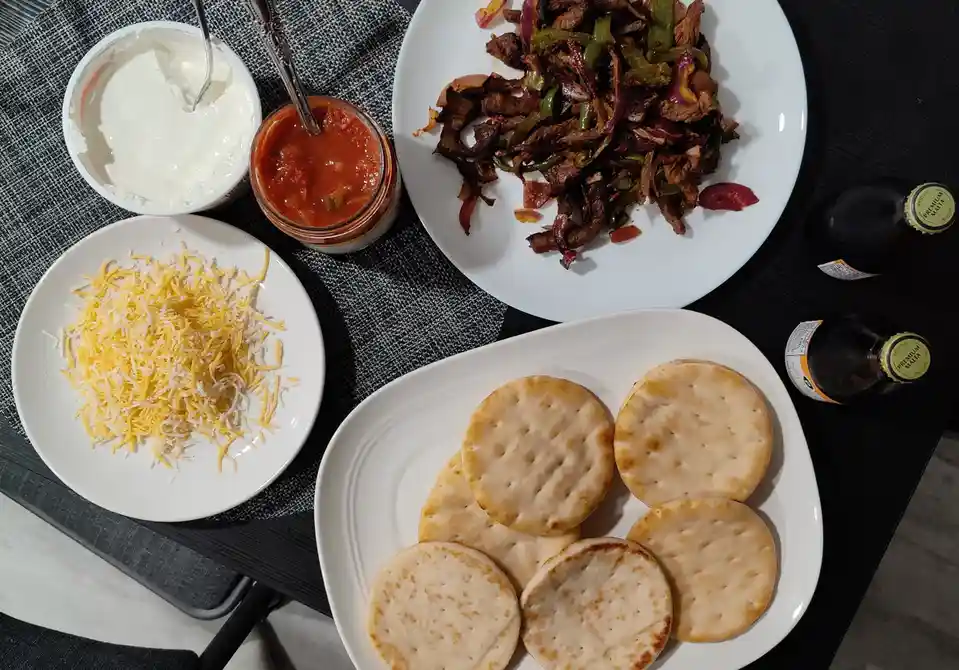 An overhead photo shows ingredients for fajitas: a plate of cooked meat and peppers, a plate of round crackers, a plate of…