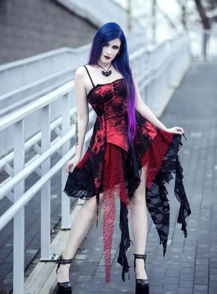 A woman with long blue and purple hair, wearing a red and black lace dress with high heels, poses on a sidewalk next to a…
