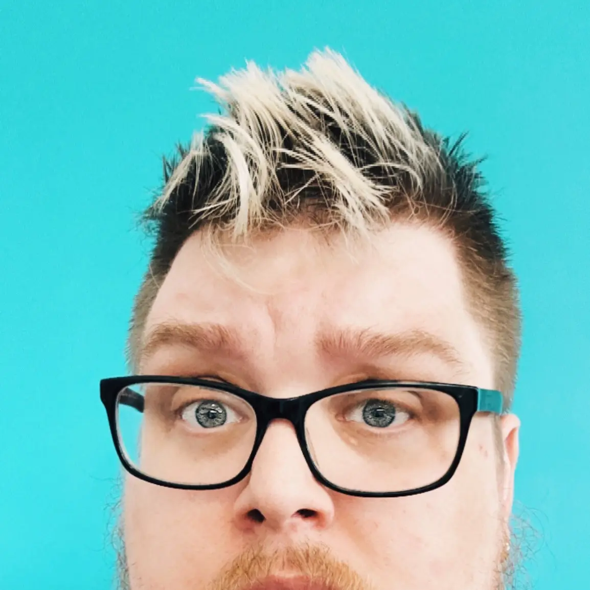 A close-up photo of a man with spiky, bleached blonde hair against a solid turquoise background, wearing black-rimmed…