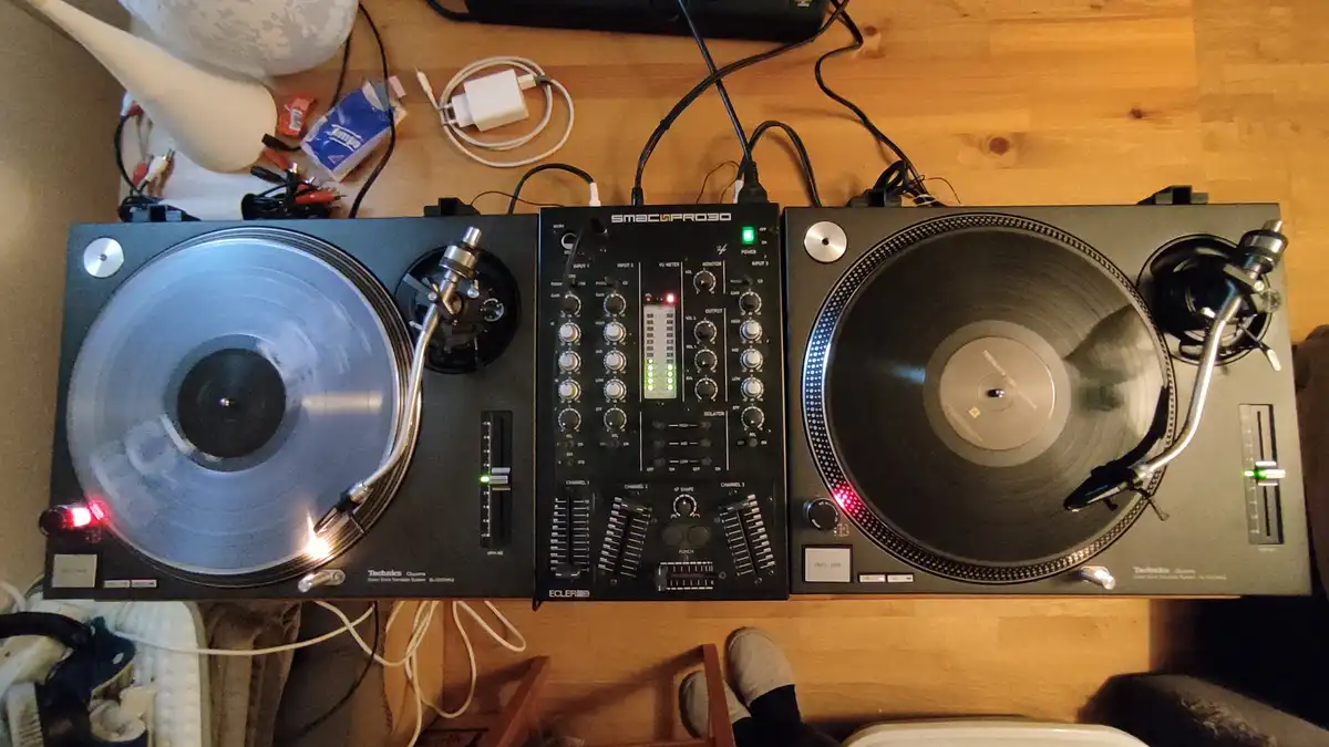 An overhead shot of a DJ setup with two Technics turntables and a central mixer on a wooden floor, surrounded by cables and…