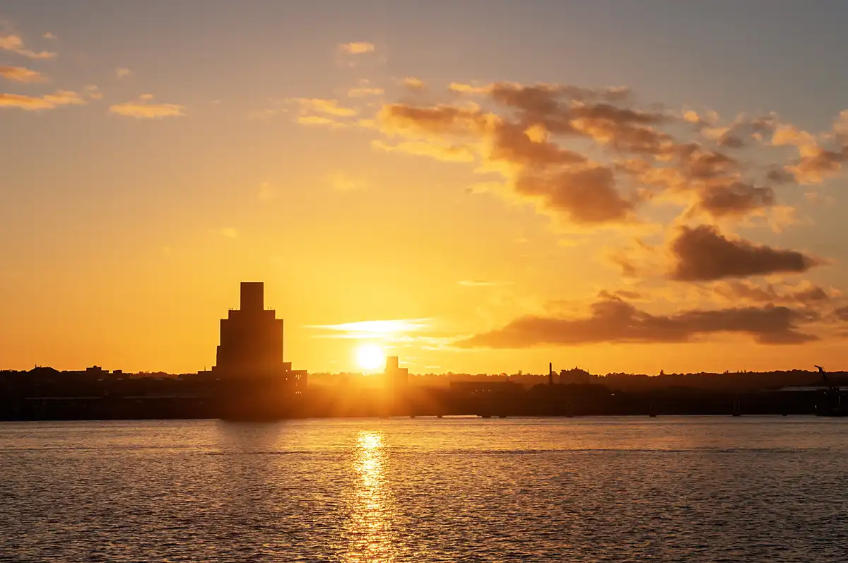 A serene sunset over a body of water with silhouetted buildings, including a prominent tower, against a sky filled with…