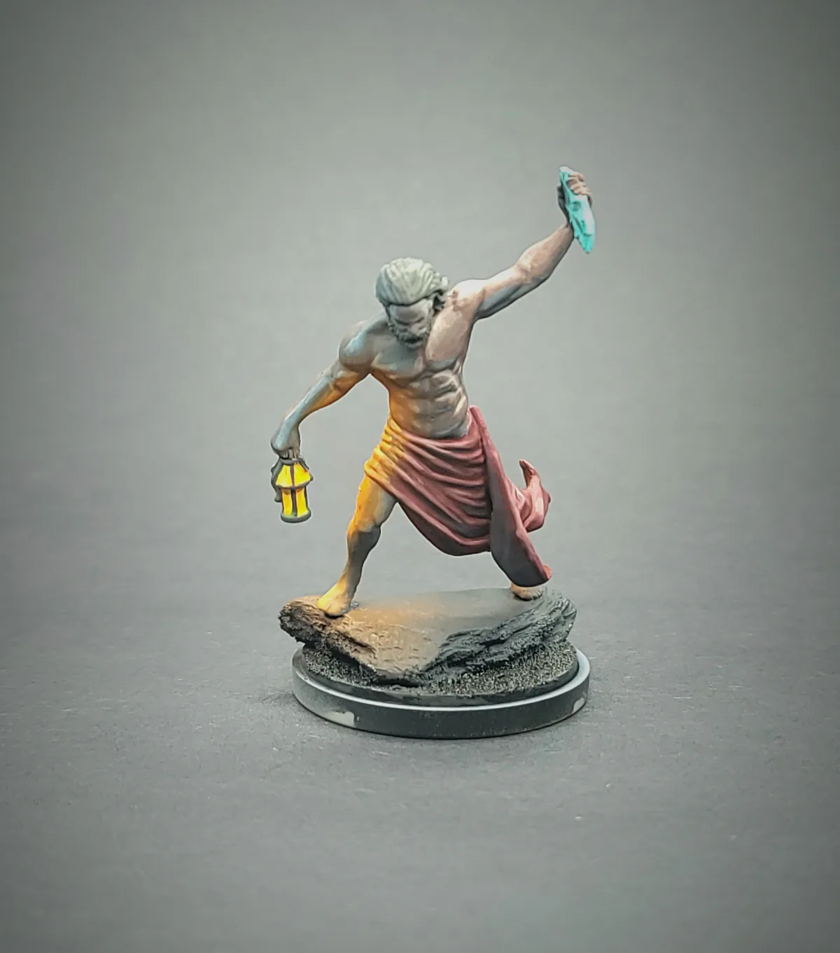 A painted tabletop miniature of a muscular, bearded man in a red loincloth stands on a rocky base, holding a glowing lantern…