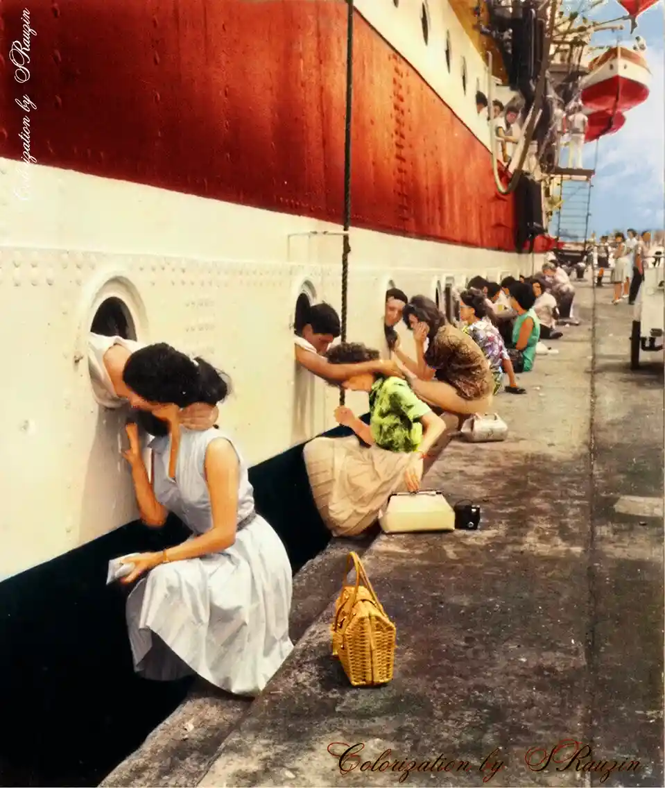 A vintage colorized photo shows a line of people sitting on a dock, each peering through portholes in the side of a large…