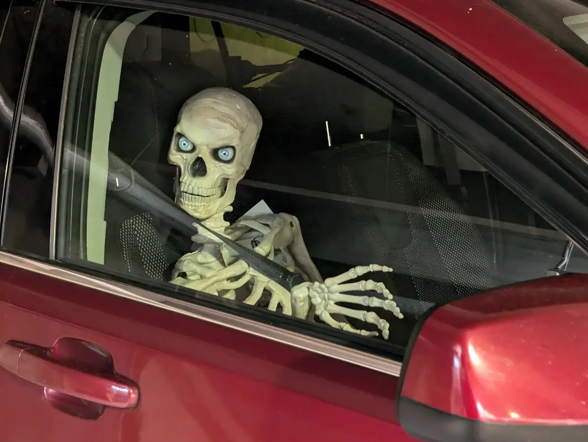 A plastic skeleton with glowing blue eyes sits in the driver's seat of a red car, wearing a seatbelt and looking out the…