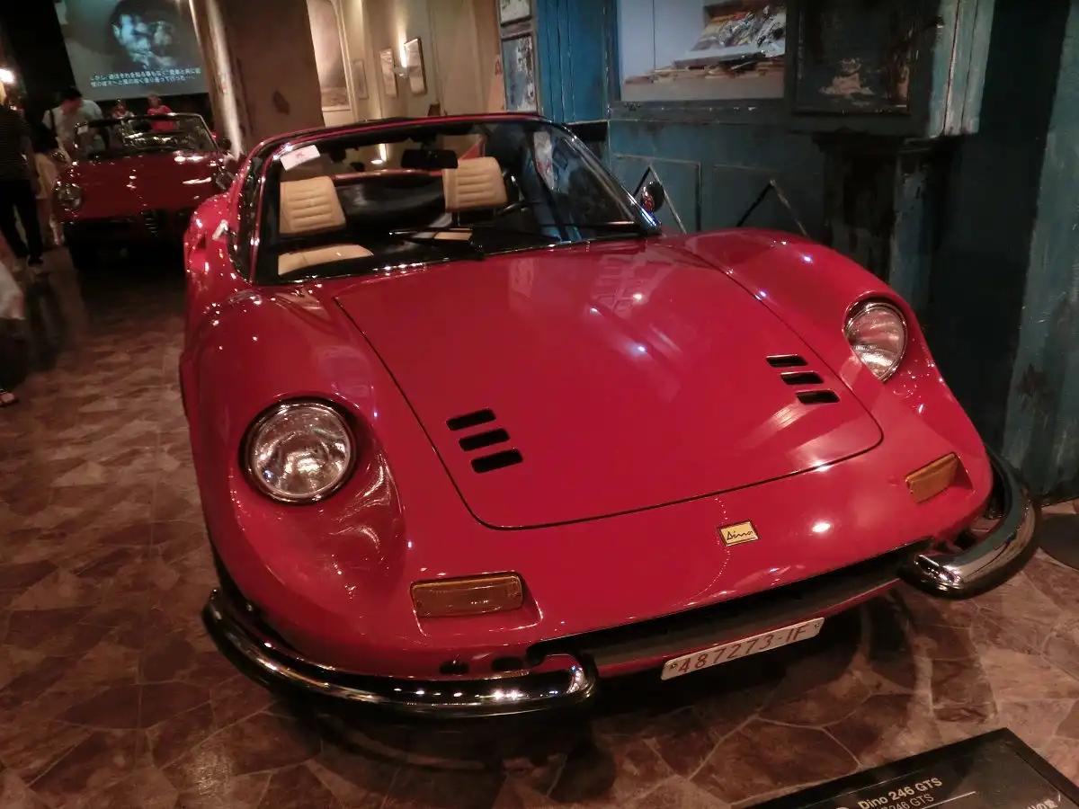 A glossy red Ferrari Dino 246 GTS is displayed indoors on a tiled floor, with its hood vents and round headlights visible…