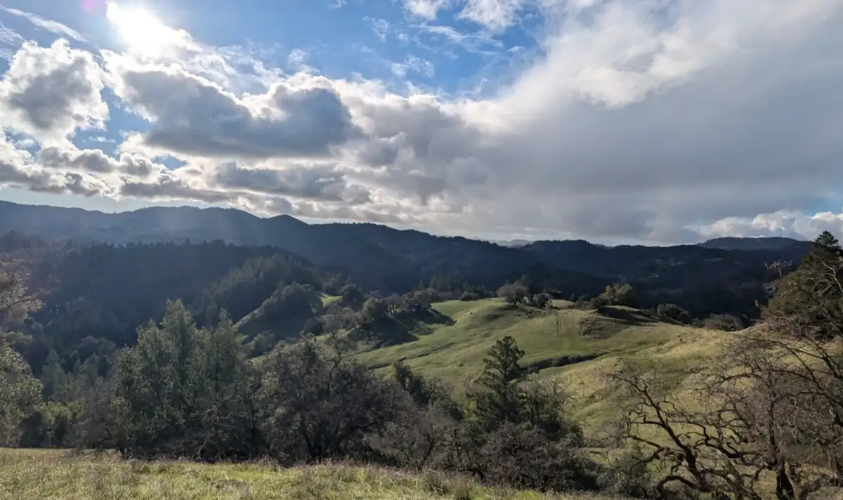 A scenic landscape photo of rolling green hills, forests, and distant mountains under a partly cloudy sky with sunlight…