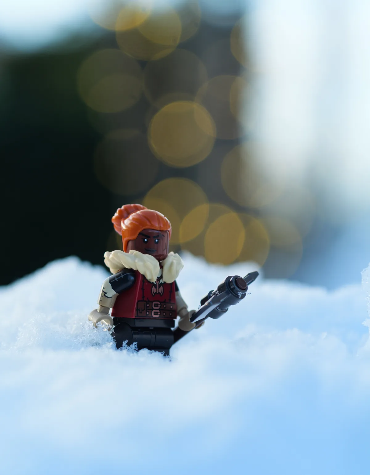 A Lego minifigure with orange hair and a red vest stands in a snowy landscape, holding a black weapon. The background…