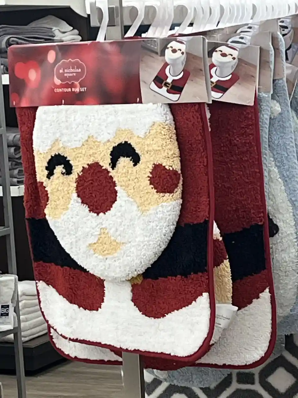A plush, red-and-white Santa Claus face rug hangs on a display rack in a store, featuring a cheerful Santa with a red nose…