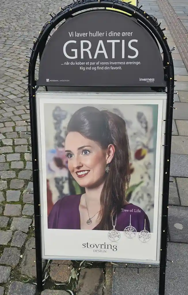A black-framed outdoor advertising sign for a jewelry store, featuring a photo of a smiling woman wearing earrings and a…