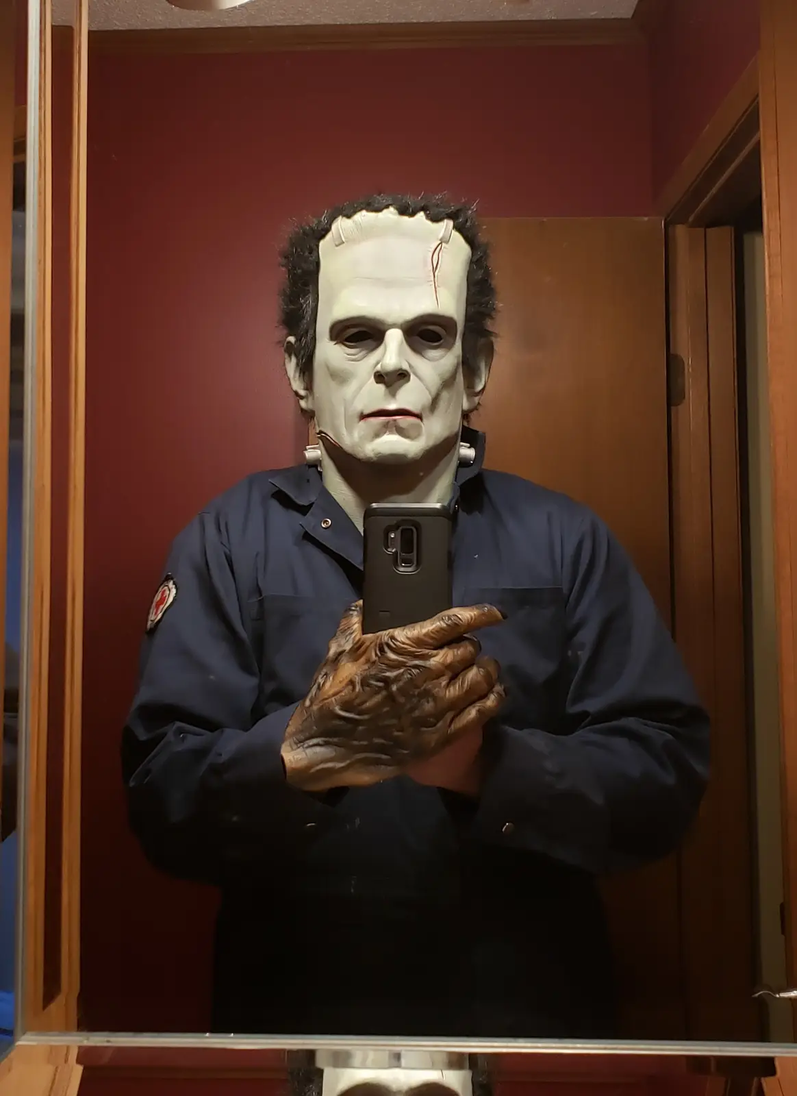 A person wearing a Frankenstein monster mask and a dark blue uniform with a red cross patch on the sleeve is taking a mirror…