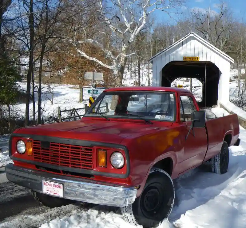 A red vintage pickup truck is parked in front of a white wooden covered bridge in a snowy, wooded area. The truck has a…