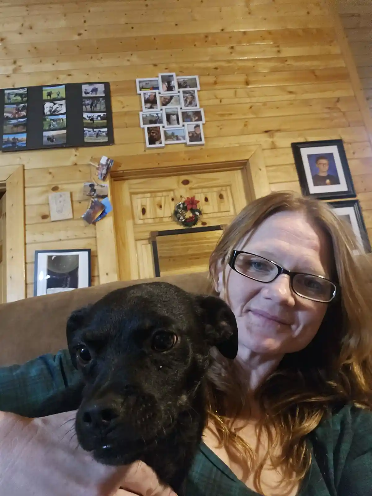 A woman with glasses smiles while holding a black dog in a cozy wooden cabin. The background shows a wood-paneled wall with…