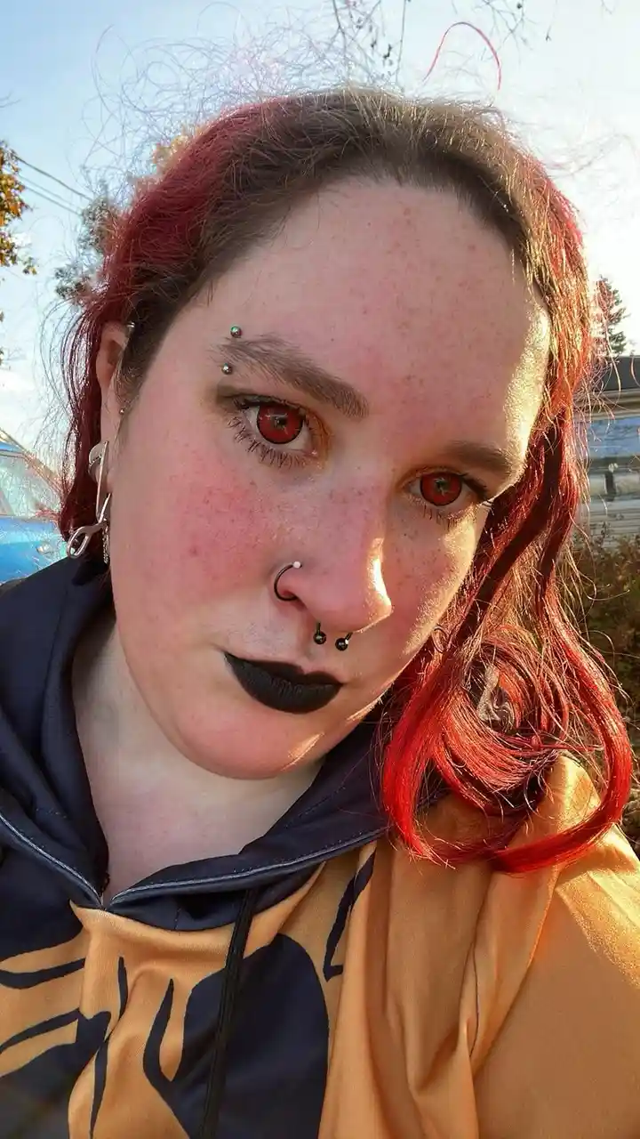 A person with red hair, red eyes, black lipstick, and multiple facial piercings stares directly at the camera. They wear a…