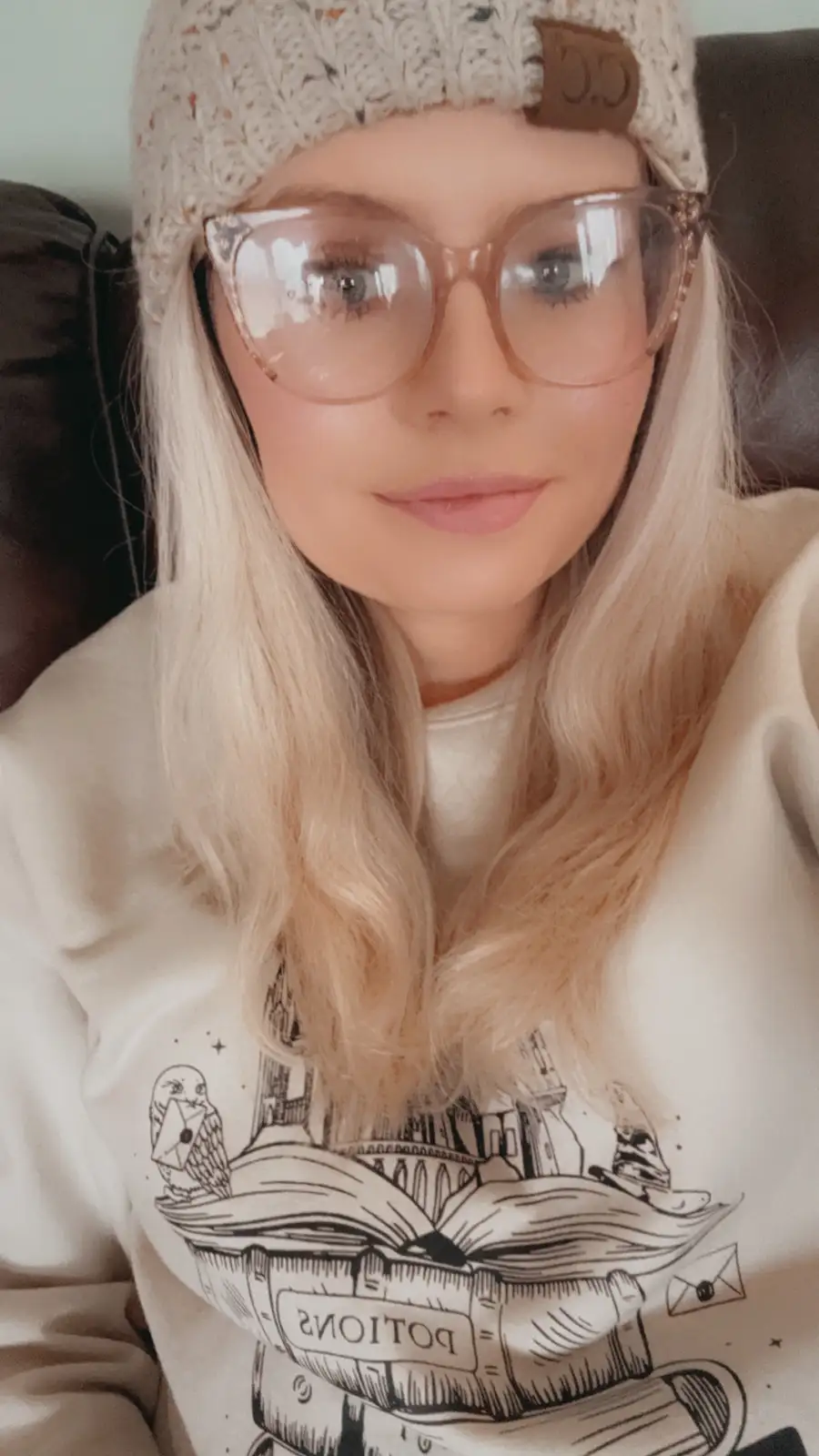 A person with blonde hair, wearing a beige knit beanie and large clear-framed glasses, looks at the camera while seated…