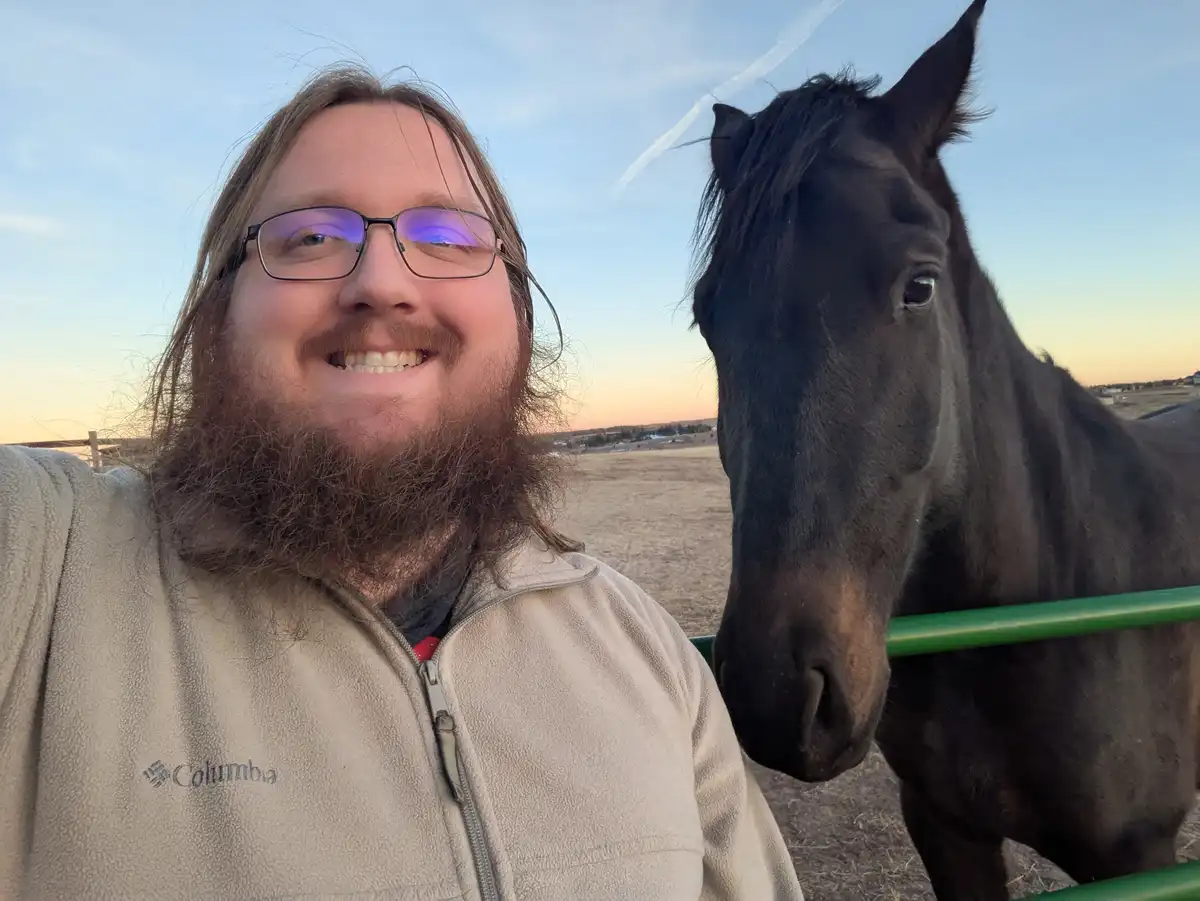 A man with glasses and a beard smiles at the camera while taking a selfie next to a black horse that is looking directly at…