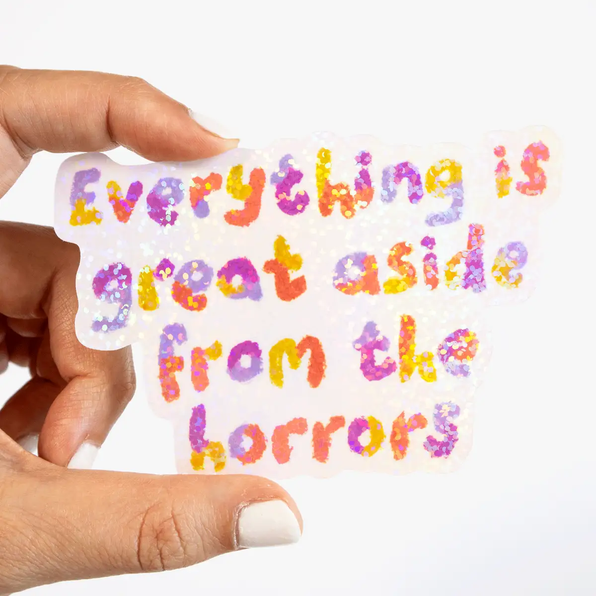 A hand with white nail polish holds a glittery, iridescent sticker with the text 'Everything is great aside from the…