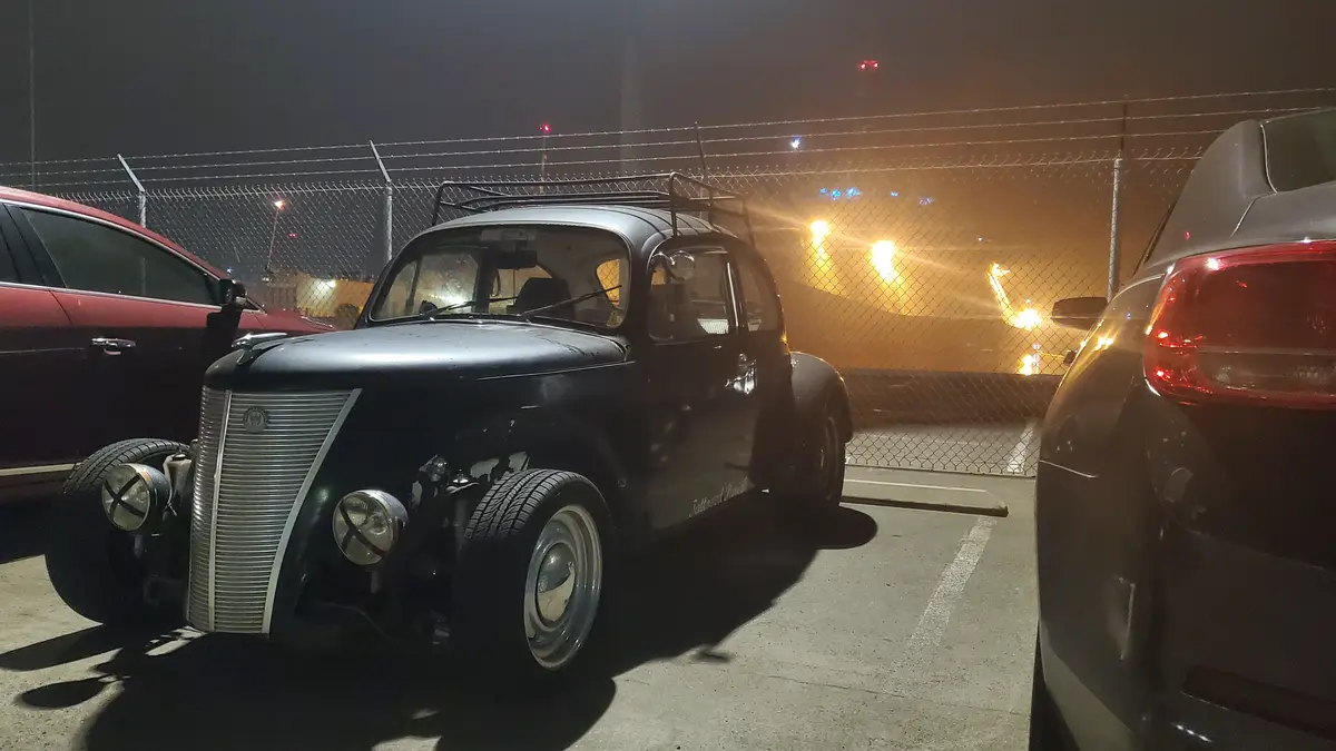 A black vintage car with a roof rack and exposed engine is parked in a dimly lit parking lot at night, flanked by a red car…