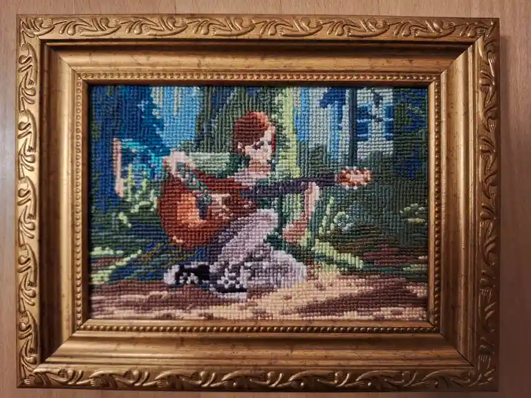 A framed needlepoint artwork depicts a pixelated character from the game 'The Legend of Zelda: Breath of the Wild' sitting…