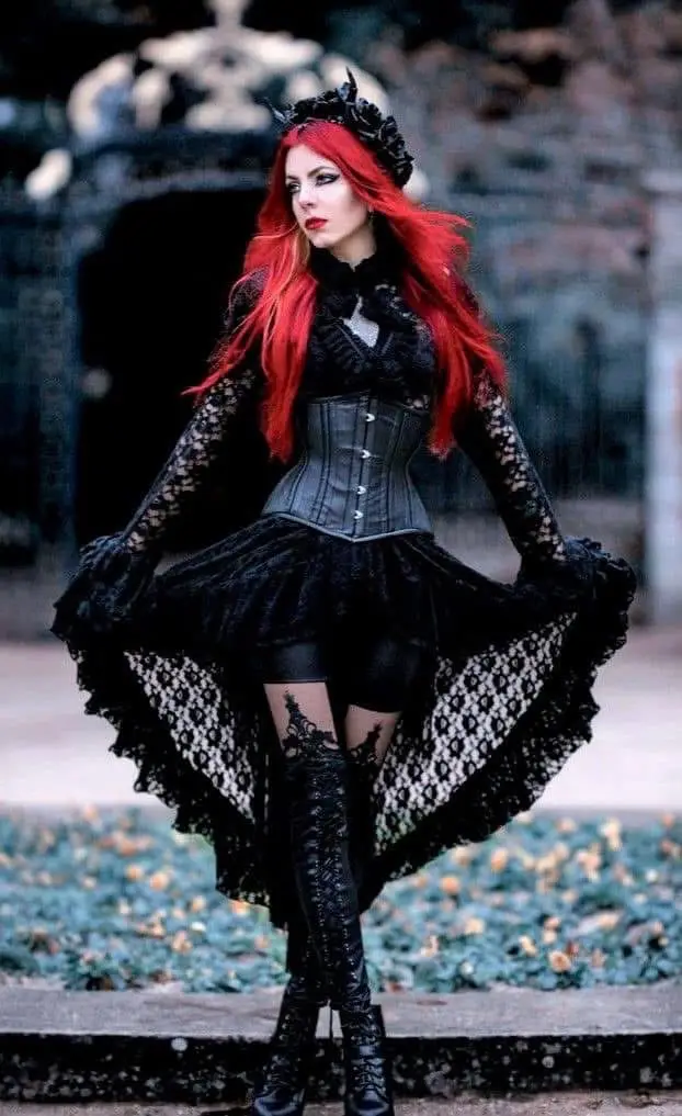 A woman with long red hair and gothic makeup poses in a black lace dress, corset, and thigh-high boots, standing outdoors in…