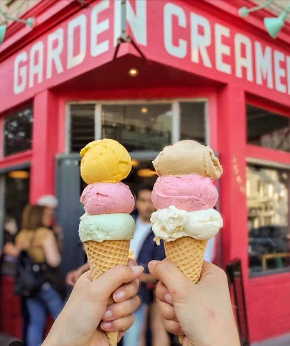Two hands hold up ice cream cones with multiple scoops each in front of a bright red building with 'GARDEN CREAMERY' written…