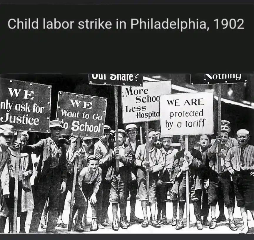 A historical black-and-white photograph shows a group of young children, likely aged 8-15, standing in a line holding…