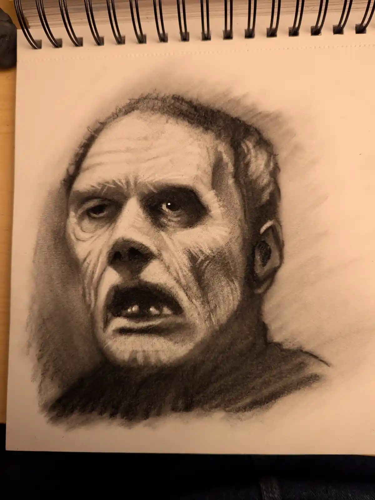 A detailed charcoal sketch on spiral-bound paper depicts a man with a distressed, screaming expression, his mouth open…