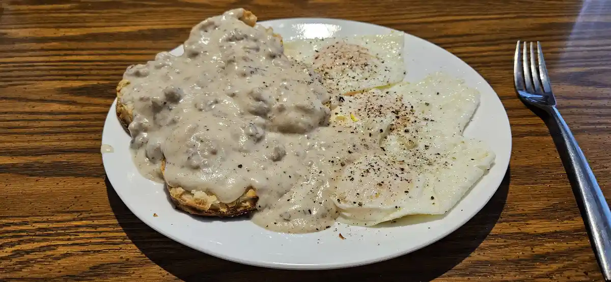 A plate of breakfast food on a wooden table, featuring biscuits smothered in creamy sausage gravy and two fried eggs…