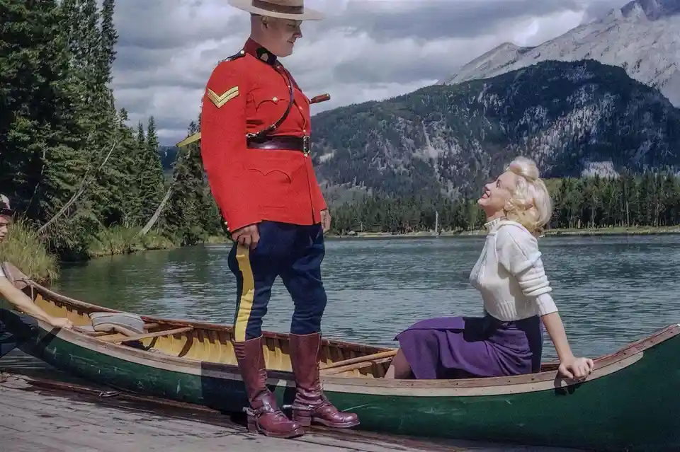 A man in a red Canadian Mountie uniform stands at the edge of a dock next to a green canoe, while a smiling blonde woman…