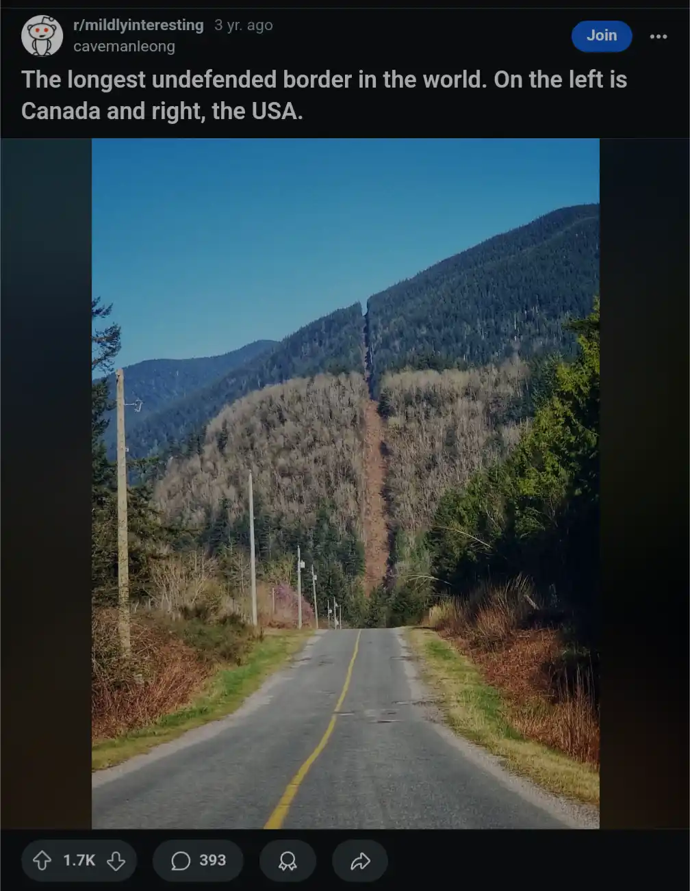A straight road runs through a forested mountain valley, with a clear, narrow, dirt-colored line splitting the hillside down…